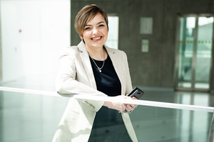 Asta Pundzienė leaning on glass railing in a bright educational facility hallway, wearing a light blazer, neutral background with corridor and windows.