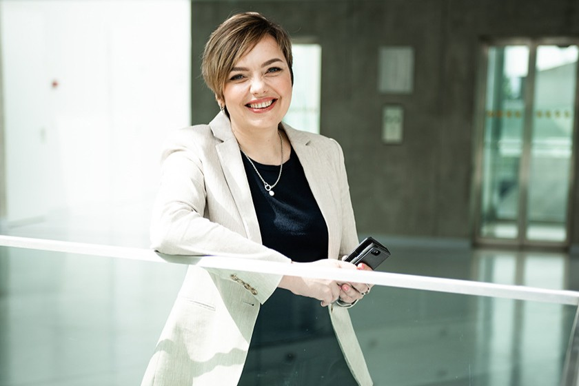 Asta Pundzienė leaning on glass railing in a bright educational facility hallway, wearing a light blazer, neutral background with corridor and windows.