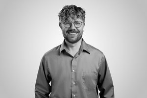 A black and white portrait of Lukas Geryba, a young man with curly hair, glasses, and casual shirt, facing camera with a neutral studio‑style background.